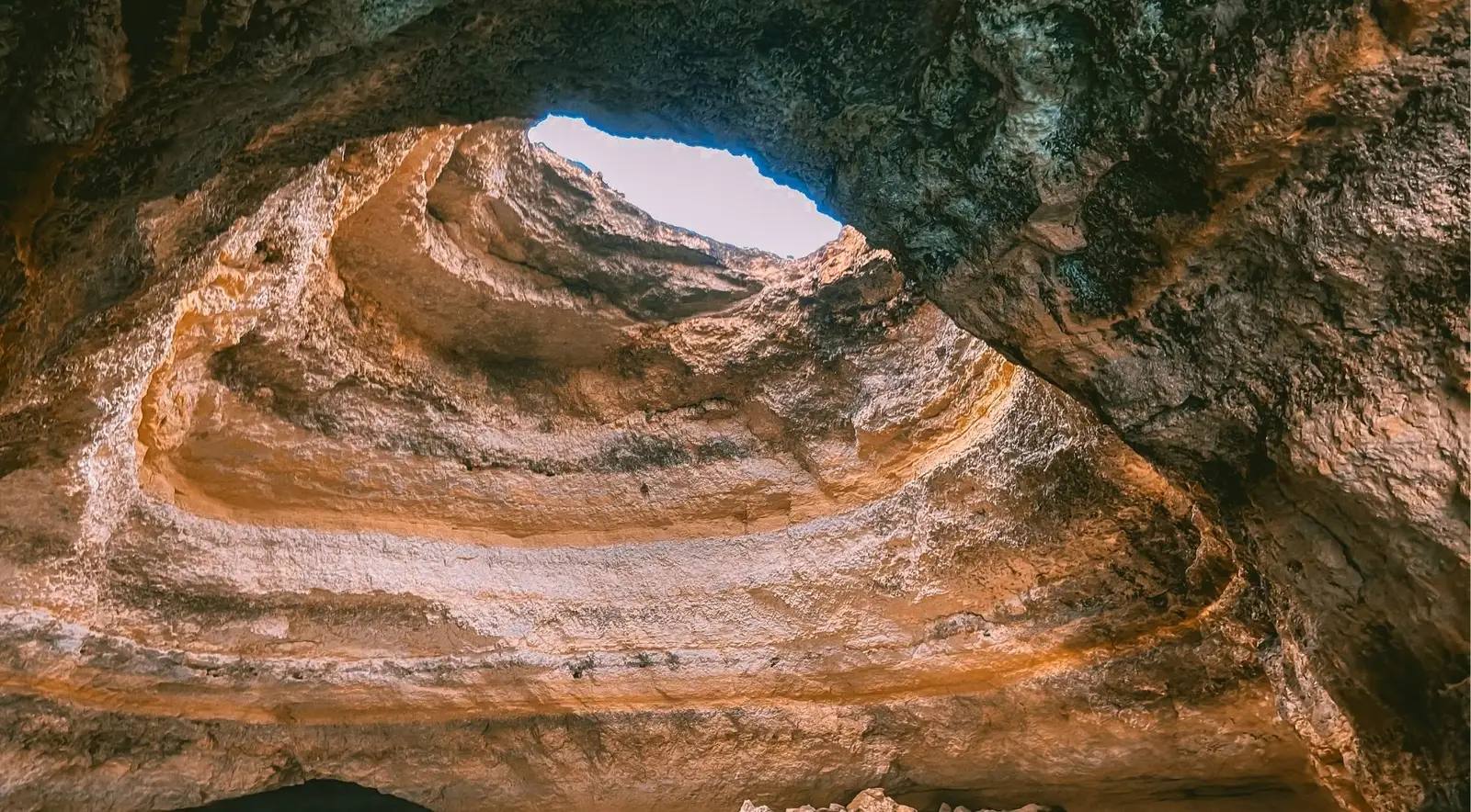 Looking up at the round skylight of Benagil Cave from inside, with blue sky above the layered golden limestone